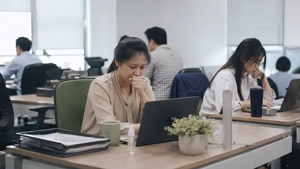 Karyawan perempuan sedang batuk dan tetap bekerja menggunakan laptop di kantor