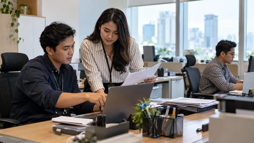 Dua rekan kerja sedang memeriksa dokumen dan mendiskusikan pekerjaan bersama di depan laptop.