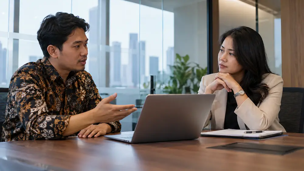 Seorang pria dan wanita sedang berdiskusi secara serius di ruang kantor dengan laptop terbuka di hadapan mereka.